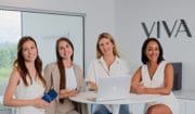 Quatre femmes sont assises à une table blanche dans un bureau moderne près d'une fenêtre, avec un ordinateur portable et un produit Clinique médico-esthétique Viva. Elles sourient, le mot VIVA étant affiché sur le mur derrière elles.