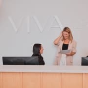 Deux femmes à la réception ; l'une assise derrière un ordinateur, souriant à l'autre, debout, tenant un presse-papiers. Le mur derrière elles affiche fièrement « VIVA », reflétant l'atmosphère chaleureuse des Cliniques Viva Québec.