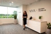 Une femme en robe noire se tient dans un bureau moderne et lumineux, doté de grandes fenêtres, de plantes d'intérieur et d'un comptoir blanc. Sur le mur, on peut lire « VIVA | bien dans ma peau.ca ».